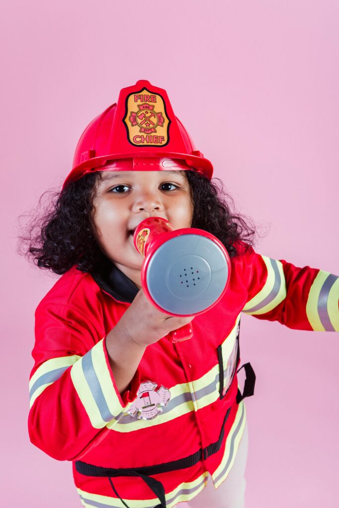 Child with curly hair in firefighter costume and helmet, playfully using a megaphone in a bright studio.